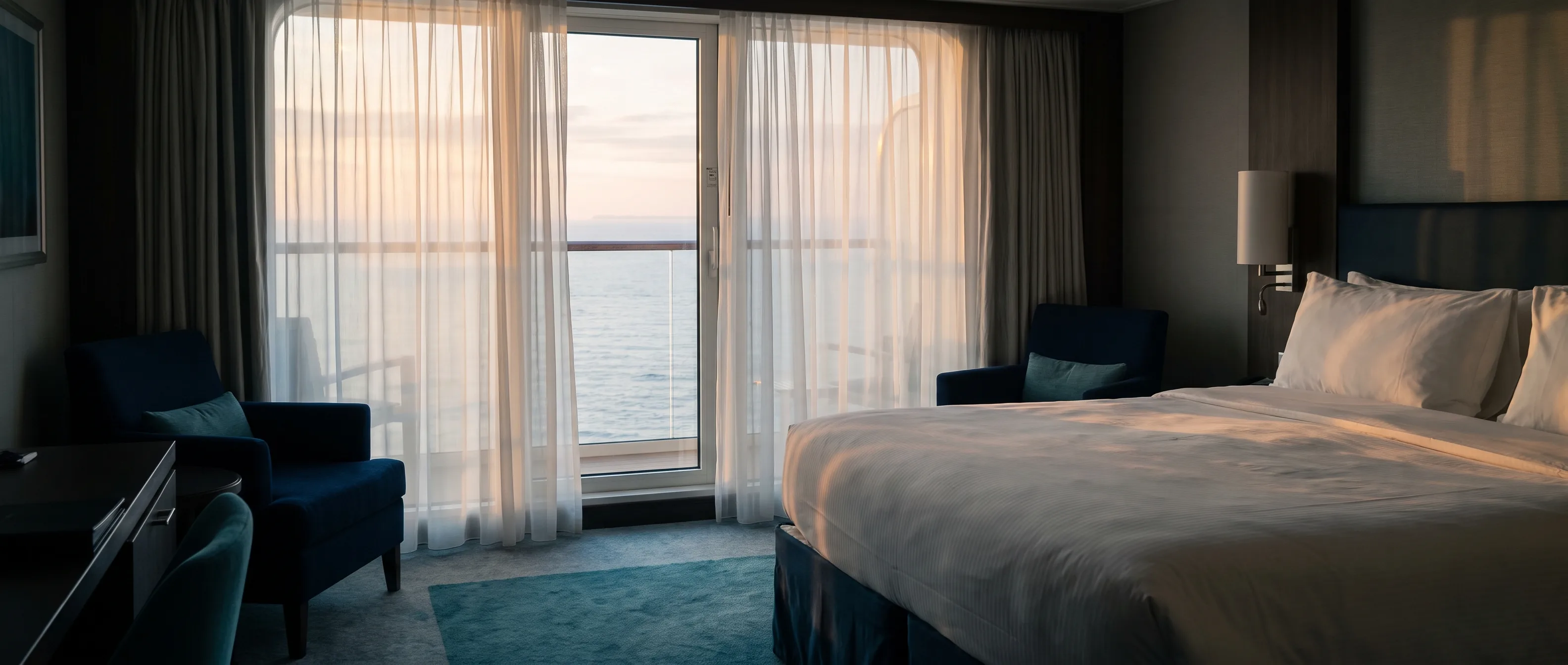 A cruise cabin at first light, sheer curtains and the calm sea beyond the window